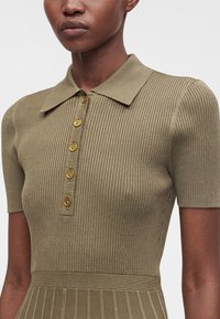 Personne à la peau foncée portant une robe polo à manches courtes en tricot côtelé vert olive avec cinq boutons dorés sur le devant.