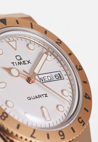 Roségoldene Timex Q Uhr mit weißem Zifferblatt, Quarzwerk, Tag/Datum-Anzeige und leuchtenden Zeigern. Sie verfügt über kantige Stundenmarkierungen und eine drehbare Lünette.