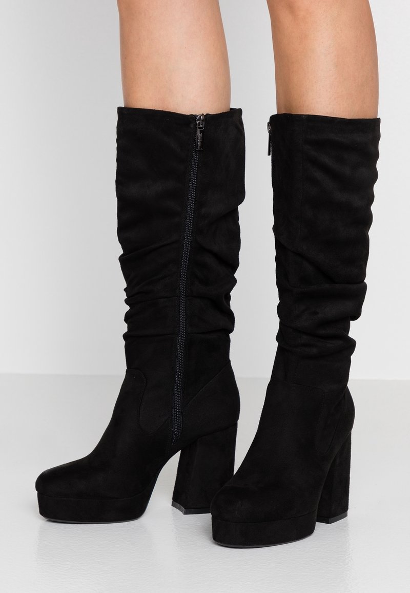 Botas negras de ante hasta la rodilla con un diseño arrugado, que cuentan con un cierre de cremallera lateral y un tacón grueso. Textura suave.