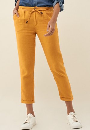 Femme portant un pantalon jaune moutarde retroussé avec une taille à cordon, une chemise bleue rentrée et des baskets blanches, debout sur un fond beige.