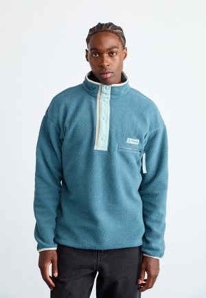 Fleece pullover in teal with beige accents, mock neck, front half-button placket, and zippered chest pocket. Soft texture and casual design.