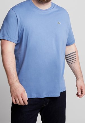 Mężczyzna w niebieskiej koszulce z krótkim rękawem z małym logo krokodyla i czarnym tatuażem w kształcie gradientowego paska na lewym przedramieniu.