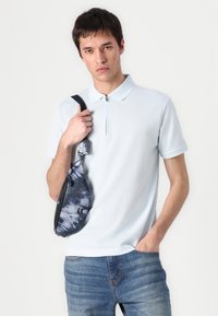 Light blue, short-sleeved polo shirt with a zippered collar. Paired with a patterned dark blue sling bag, worn over a pair of denim jeans.