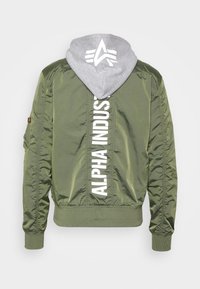 Oliivinvihreä pommikaukarjakkua, jossa on harmaa huppari kiinnitettynä. Takana on valkoinen "ALPHA INDUSTRIES" -logo pystysuoraan ja hihansuut ovat joustavat.