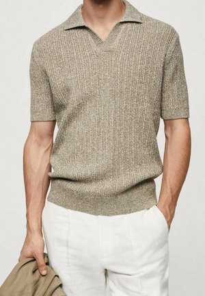 Mand iført beige kortærmet strikket polo-sweater og hvide bukser, der holder en beige jakke i venstre hånd i taljehøjde.