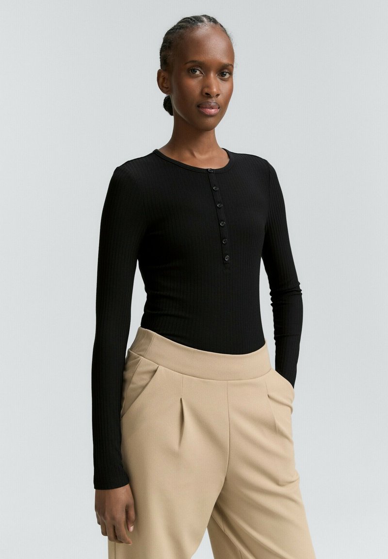 Zwart geribbeld langemouwen top met knoopdetail bij de kraag, gecombineerd met beige hoge taille broek met plooidetails.