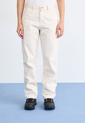 Pantaloni beige a righe sottili con una vestibilità rilassata, caratterizzati da righe rosse, tasche anteriori e chiusura con bottone. Abbinati a scarpe nere con lacci.