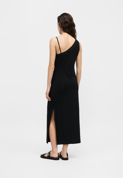 Γυναίκα με μαύρο μίντι φόρεμα ενός ώμου με πλαϊνή σχισμή και μαύρα φλατ σανδάλια, που στέκεται μπροστά σε απλό φόντο.