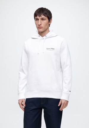 Młody mężczyzna ubrany w biały kaptur Tommy Hilfiger z przednią kieszenią i ciemnoniebieskie dżinsy, stojący na jednolitym jasnym tle.