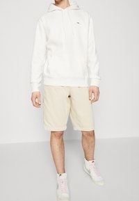 Persona con una sudadera blanca, pantalones cortos beige y zapatillas altas blancas de Nike con el swoosh verde, de pie frente a un fondo liso claro.