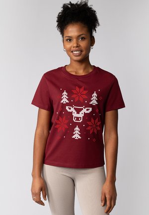 Jeune femme souriante, portant un t-shirt bordeaux avec des rennes, des flocons de neige et des sapins de Noël en pixels blancs et rouges, se tenant devant un fond uni.