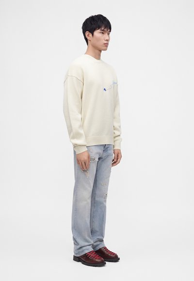 Jeune homme portant un pull crème, un jean clair déchiré et des bottes marron foncé avec des lacets rouges, debout devant un fond blanc uni.