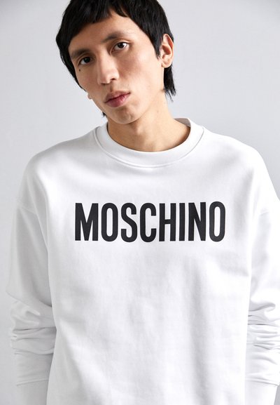 Hvit genser laget av bomullsblanding, med en fremtredende svart "MOSCHINO" logo foran. Rund hals og lange ermer.