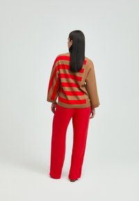 Brauner Strickpullover mit roten horizontalen Streifen, ausgestattet mit weiten Ärmeln. Kombiniert mit lockeren roten Hosen. Einheitliche Farbe und Textur.