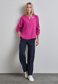 Blusa in cotone rosa a maniche lunghe con scollatura a V e chiusura con bottoni, abbinata a pantaloni a gamba larga navy e sneaker bianche. Texture liscia.