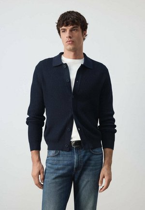 Joven con un cárdigan de punto azul marino con botones encima de una camiseta blanca y vaqueros azules, de pie frente a un fondo claro y liso.