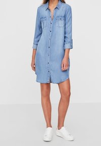 Jeanskleid, hellblau, mit durchgehender Knopfleiste, zwei Brusttaschen, aufgekrempelte Ärmel, knielang, kombiniert mit weißen Turnschuhen.