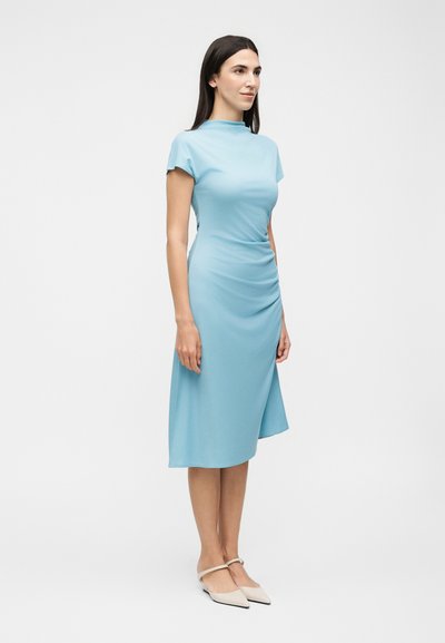 Γυναίκα που φορά ένα ανοιχτό μπλε μίντι φόρεμα με κοντά μανίκια και λεπτομέρεια με σούρες, φορώντας λευκές πλατφόρμες μυτερές με λουράκια στον αστράγαλο.