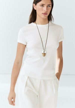 Giovane donna che indossa una semplice t-shirt bianca e pantaloni bianchi con una lunga collana con un ciondolo argentato e marrone.