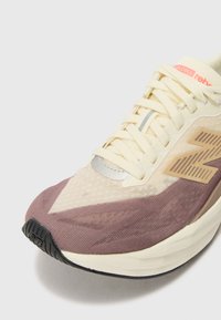 Chaussure de sport avec une tige crème, accents violets, texture en maille respirante, semelle à coussinage prononcé et détails de logo latéraux marquants.