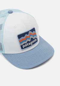 Balta un gaiši zila cepure ar platu mali, un sietiņu aizmugurē, kā arī taisnstūra izšuvums ar Patagonia logo, kuram ir krāsaini viļņveida svītras.