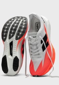 Scarpe sportive con tomaia in mesh bianco con dettagli neri e suola arancione. La suola presenta un battistrada nero testurizzato per una migliore aderenza.