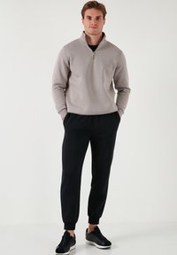 Hellgrauer Reißverschluss-Pullover mit hohem Kragen, kombiniert mit schwarzen Jogginghosen. Schwarze Sneakers mit weißen Sohlen und minimalistischem Design.