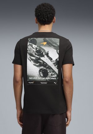 Mand iført sort t-shirt med sort-hvidt racerbilsmotiv og teksten "NEVER STOP RACING" på ryggen mod en grå baggrund.