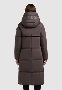 Pikk pruun puffer-kate suure kapuutsiga, millel on tikandmuster, KHUJO logo seljal ja külgsuundade lõhed liikumisvabaduse jaoks.