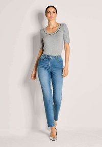 Frau in grauem Kurzarmoberteil mit perlenbesetztem Ausschnitt, hochgeschnittenen blauen Jeans und silbernen spitzen Absätzen, die vor einer schlichten Wand steht.