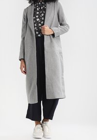 Szary długi płaszcz wykonany z teksturowanej tkaniny, z otwartym przodem, długimi rękawami i dwiema bocznymi kieszeniami, w połączeniu z czarną bluzką w wzór i ciemnymi spodniami.