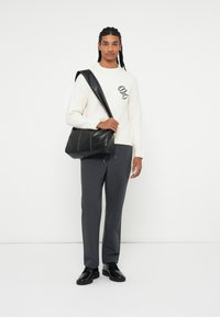 Borsa a messenger in pelle nera con tracolla regolabile, caratterizzata da cuciture bianche. Il modello indossa un maglione color crema con logo e pantaloni grigi.