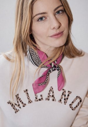 Mladá žena se světlými vlasy nosí bílou mikinu s nápisem "MILANO" v leopardím vzoru a kolem krku má uvázaný růžový šátek s leopardím potiskem.