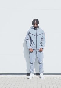 Γκρι αθλητική φόρμα από ελαφρύ υλικό, η οποία περιλαμβάνει ζακέτα με φερμουάρ και ψηλό γιακά, ταιριαστά παντελόνια και λευκά αθλητικά παπούτσια.