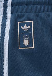 Μπλε υφασμάτινο σορτς με λευκές κάθετες ρίγες και μπεζ λογότυπο Adidas trefoil πάνω από το σήμα της ιταλικής ποδοσφαιρικής ομάδας.