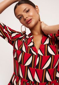 Czerwona i beżowa sukienka w wzory z dekoltem w serek i rękawami do trzech ćwiartek, z geometrycznymi kształtami i rozkloszowanym dołem. Złote kolczyki w kształcie kół.