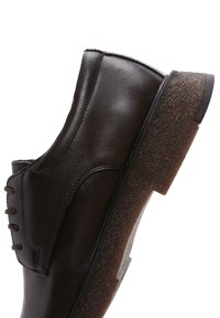 Chaussure en cuir marron avec une finition lisse, bout rond et semelle en caoutchouc marron texturée. Présente des coutures visibles et un design de lacets à cinq œillets.