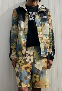 Kurtka w kwiatowy wzór w odcieniach niebieskiego, zielonego i brązowego, zestawiona z dopasowanymi szortami. Czarne akcenty i logotypy na obu elementach.