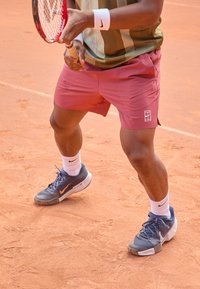 Tennisspiller i en mønstret grønn og gull skjorte, burgunder shortser og grå Nike-sko med oransje detaljer, på en leiroverflate.