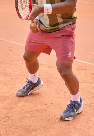 Tennis player in a patterned green and gold shirt, maroon shorts, and gray Nike shoes with orange accents, on a clay court surface.