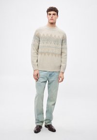 Jasnobeżowy sweter dziany z niebieskimi geometrycznymi wzorami, okrągłym dekoltem, ściągaczami przy mankietach i u dołu. Połączony z jasnoniebieskimi dżinsami i ciemnymi butami.