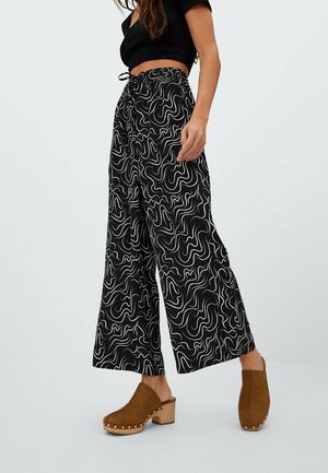 Vrouw draagt zwarte wijde broek met witte abstracte spiralen, een zwart cropped top en bruine houten klompen met gespen met studs.