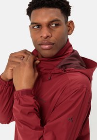 Červená nepromokavá bunda s kapucí, vybavená elastickými manžetami, detaily na zip a lehkým texturovaným materiálem. Logo na hrudi a rukávu.