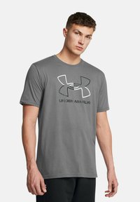 Grå kortærmet t-shirt lavet af blødt stof, med et stort sort og hvidt Under Armour-logo på forsiden. Simpelt design og rund hals.