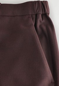 Gros plan d’un pantalon marron foncé texturé avec une ceinture élastique et une poche latérale sur un fond clair.