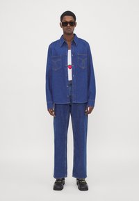 Camicia di denim blu con cuciture in stile western, indossata sopra un top bianco con una grafica di fragola, abbinata a jeans blu e scarpe nere.