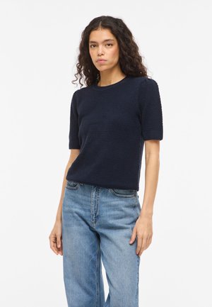 Žena s kučeravými vlasmi, ktorá má na sebe námornícky modrý krátky sveter s textúrou a svetlomodré rifle s vysokým pásom, na bielom pozadí.