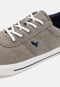 Baskets en toile grises avec des lacets blancs, oeillets gris et logo d'oiseau brodé en bleu marine. Semelle extérieure en caoutchouc blanc avec une bande bleu marine autour du bord.