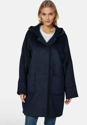 Vrouw draagt een loszittende donkerblauwe jas met capuchon, grote voorzakken en wijde mouwen over een wit topje en blauwe jeans.