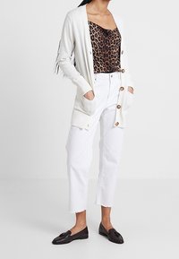 Cárdigan ligero de crema con botones y bolsillos, combinado con una camiseta de leopardo y pantalones cortos blancos. Mocasines negros completan el look.